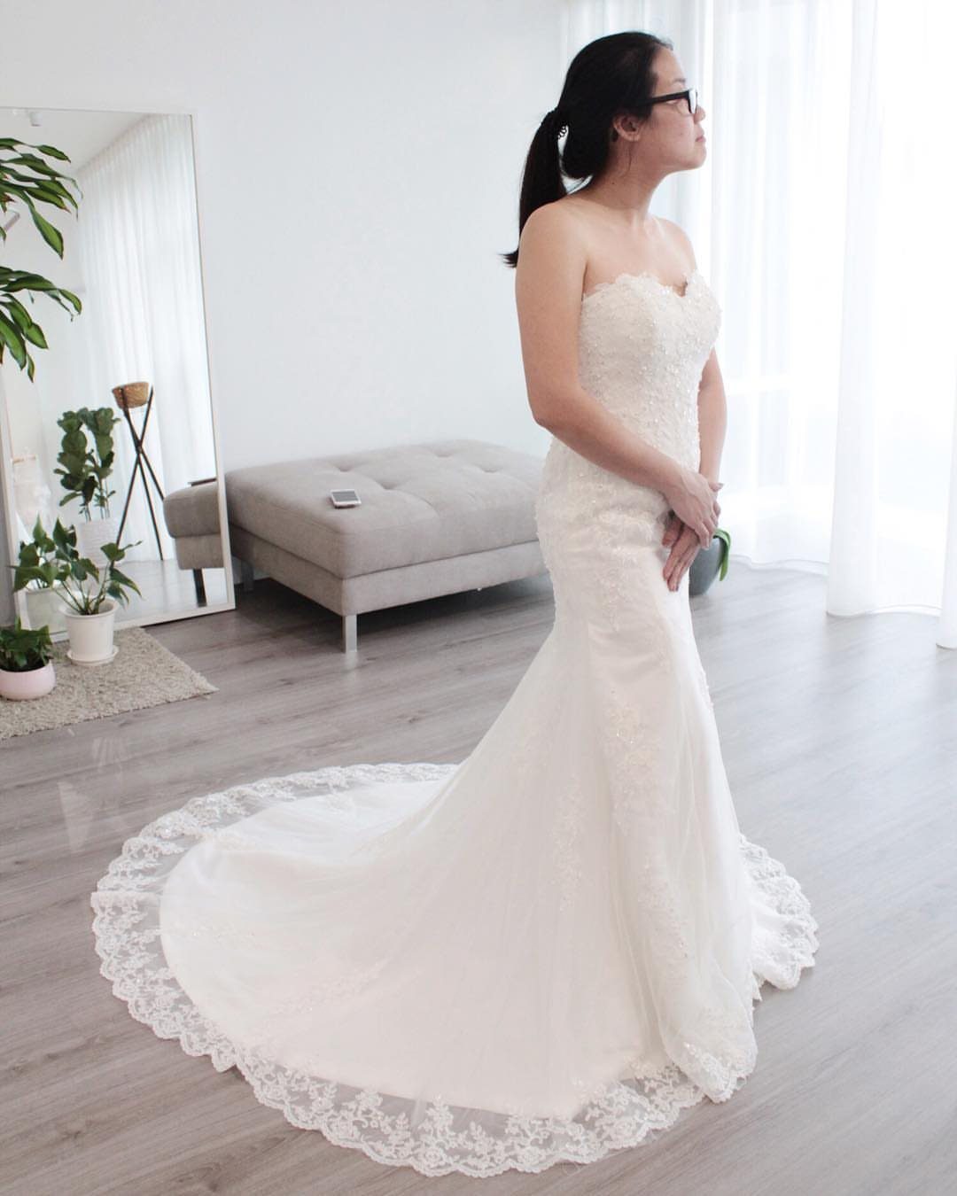A woman in a white lace strapless wedding dress with a long train standing in a bright room.