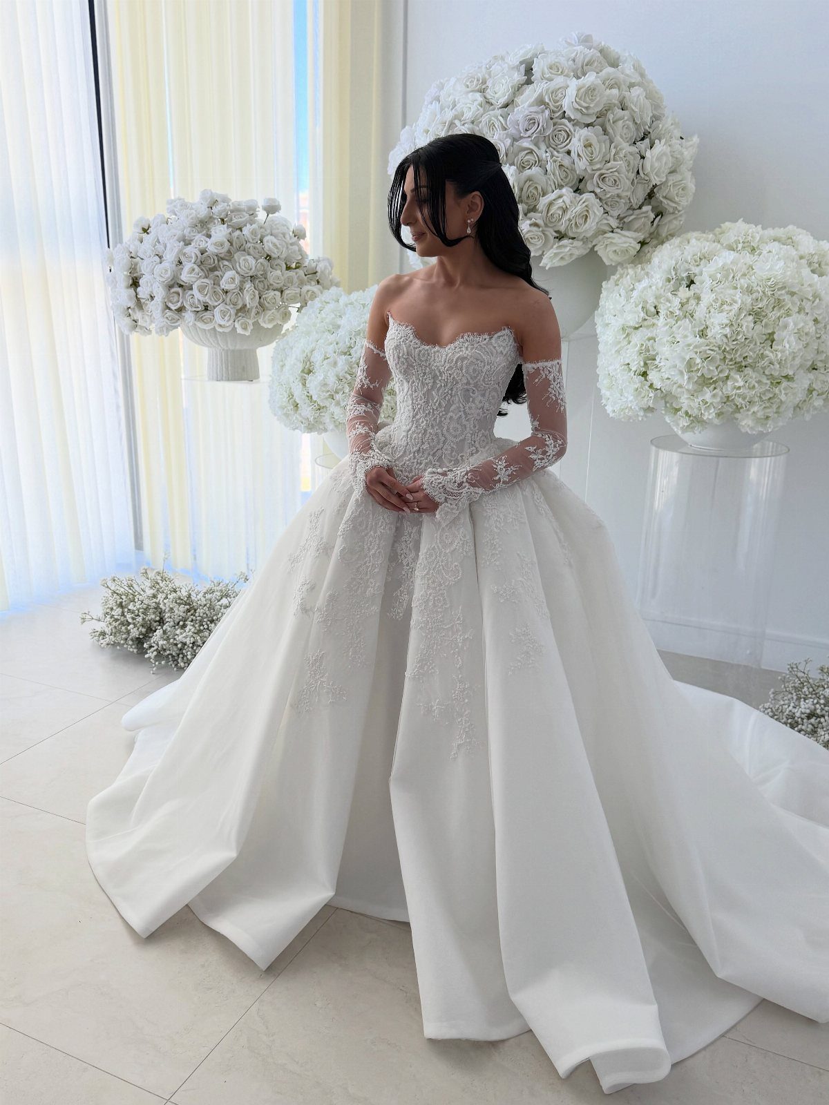 A woman wearing a white lace off-the-shoulder ball gown sitting among white flowers.