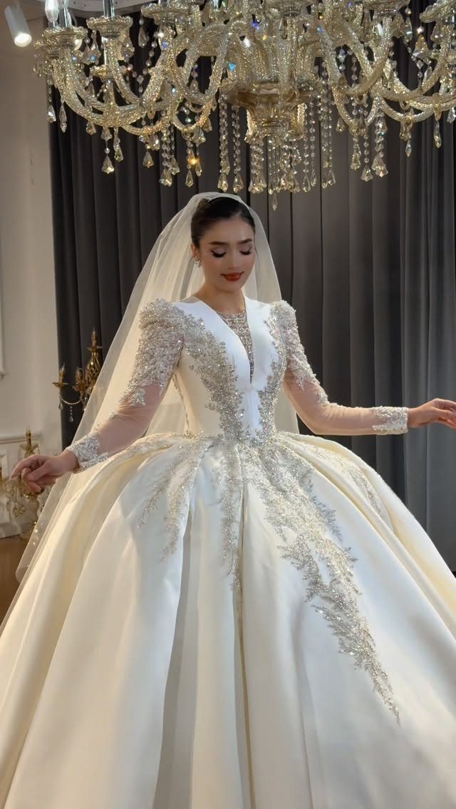 Woman in a voluminous white satin wedding gown with silver embroidery and long lace sleeves.
