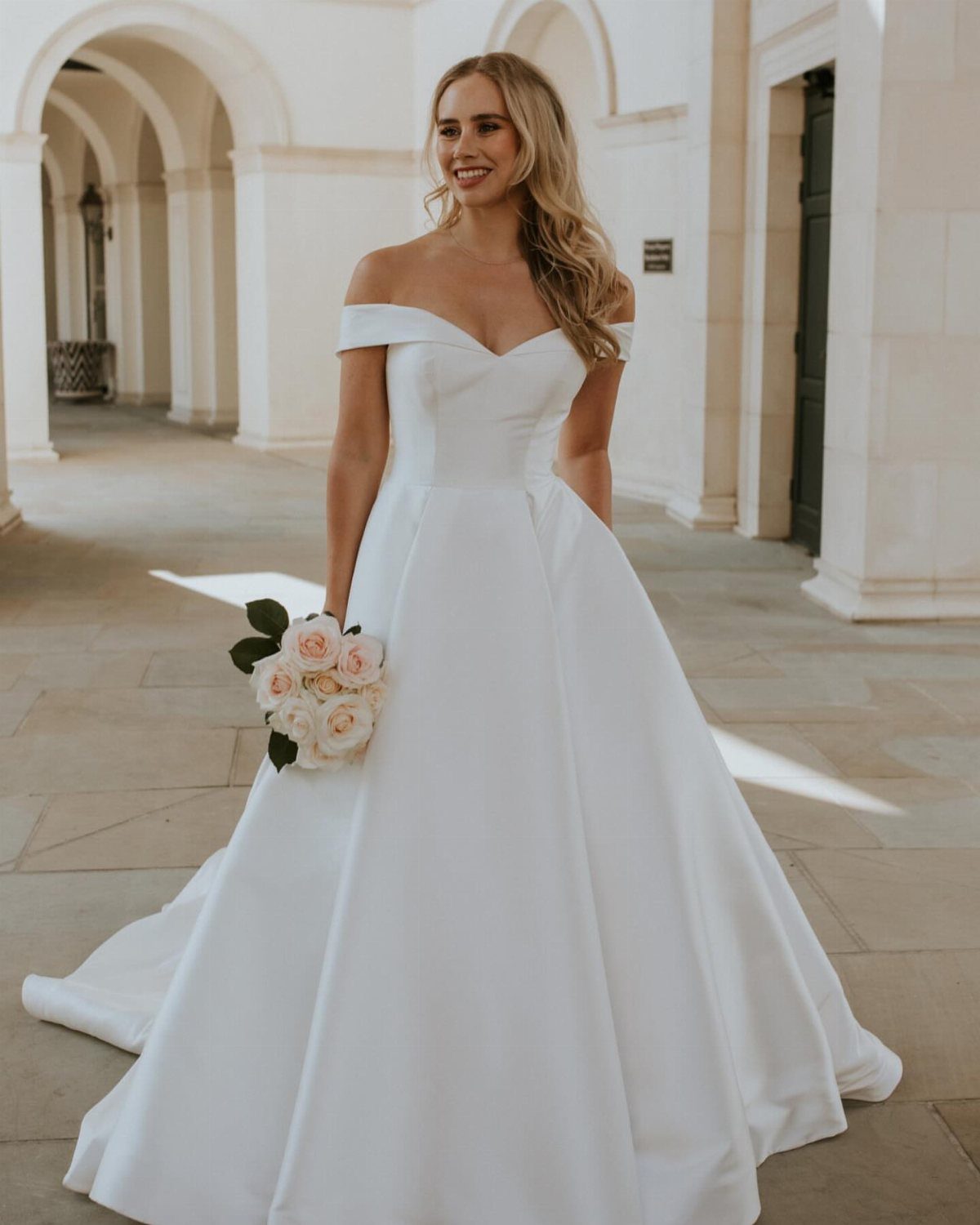 A smiling blonde bride in a white off-the-shoulder satin wedding dress holding roses.