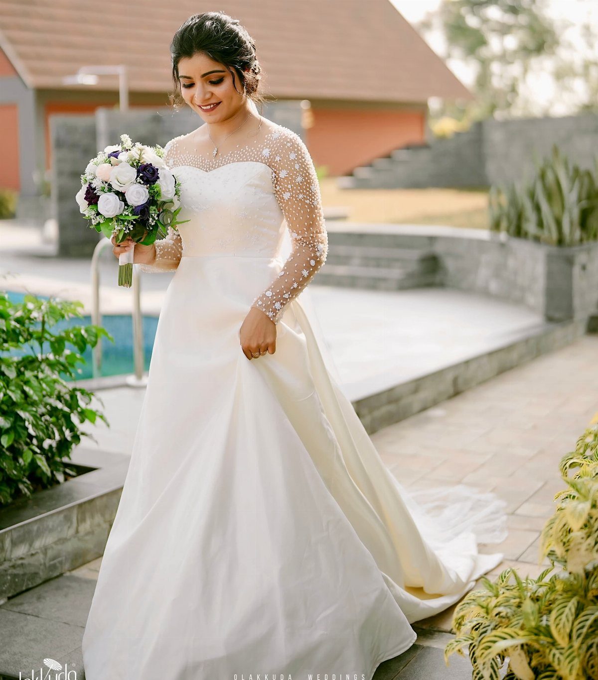A bride in a white wedding dress with pearl-embellished sheer sleeves holding a bouquet.