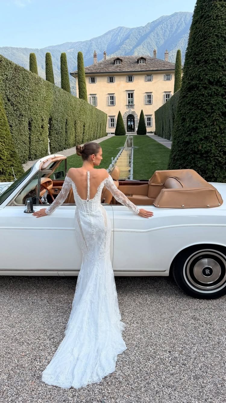 Woman in a white lace wedding dress standing beside a vintage white convertible car.