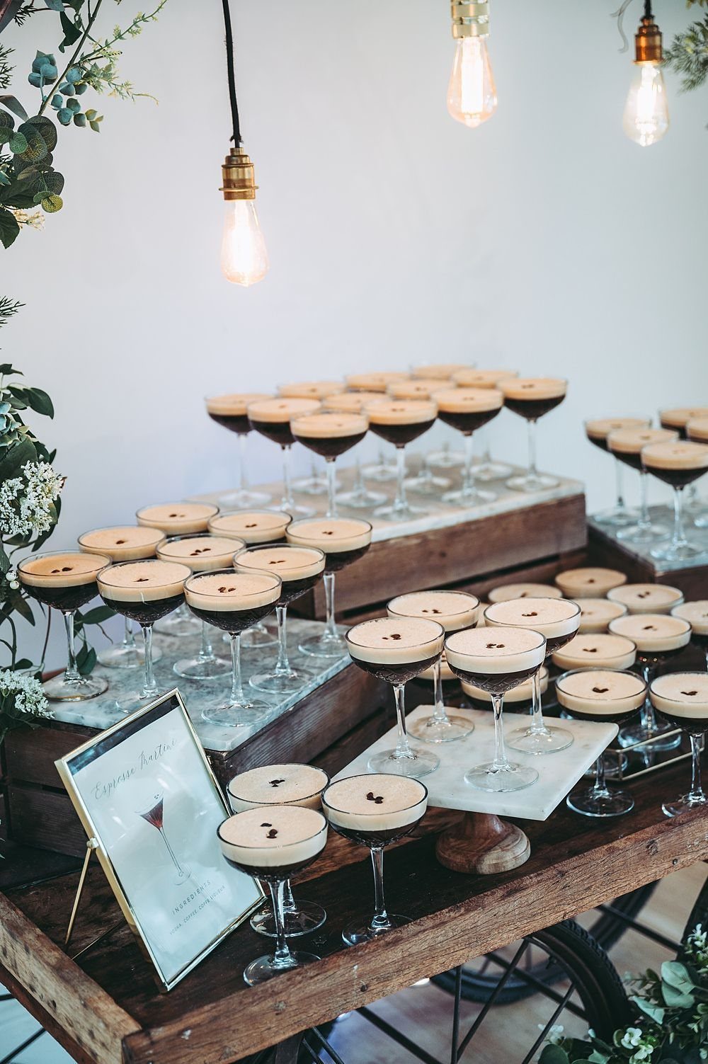 Many dark cocktails in coupe glasses arranged on wooden crates under hanging Edison bulbs.