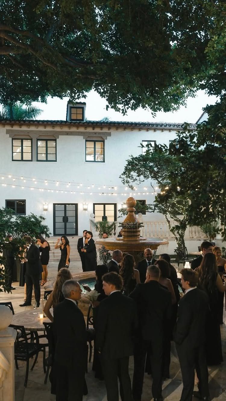 Guests dressed in formal black clothing gather in a courtyard with a fountain at dusk.