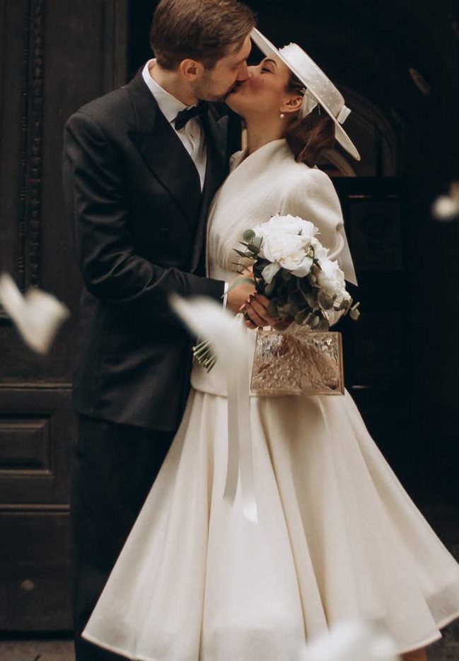 A bride in a vintage white satin dress and hat kisses her groom outdoors.