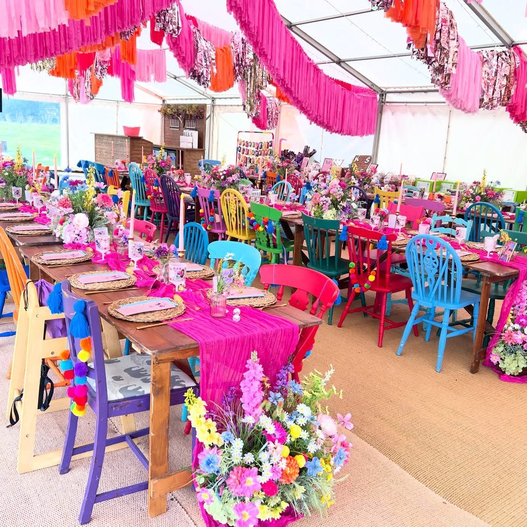 21 Colorful Wedding Table Settings for Every Style and Budget 9 Brightly colored wooden chairs surround long wooden tables decorated with pink runners and vibrant floral centerpieces.