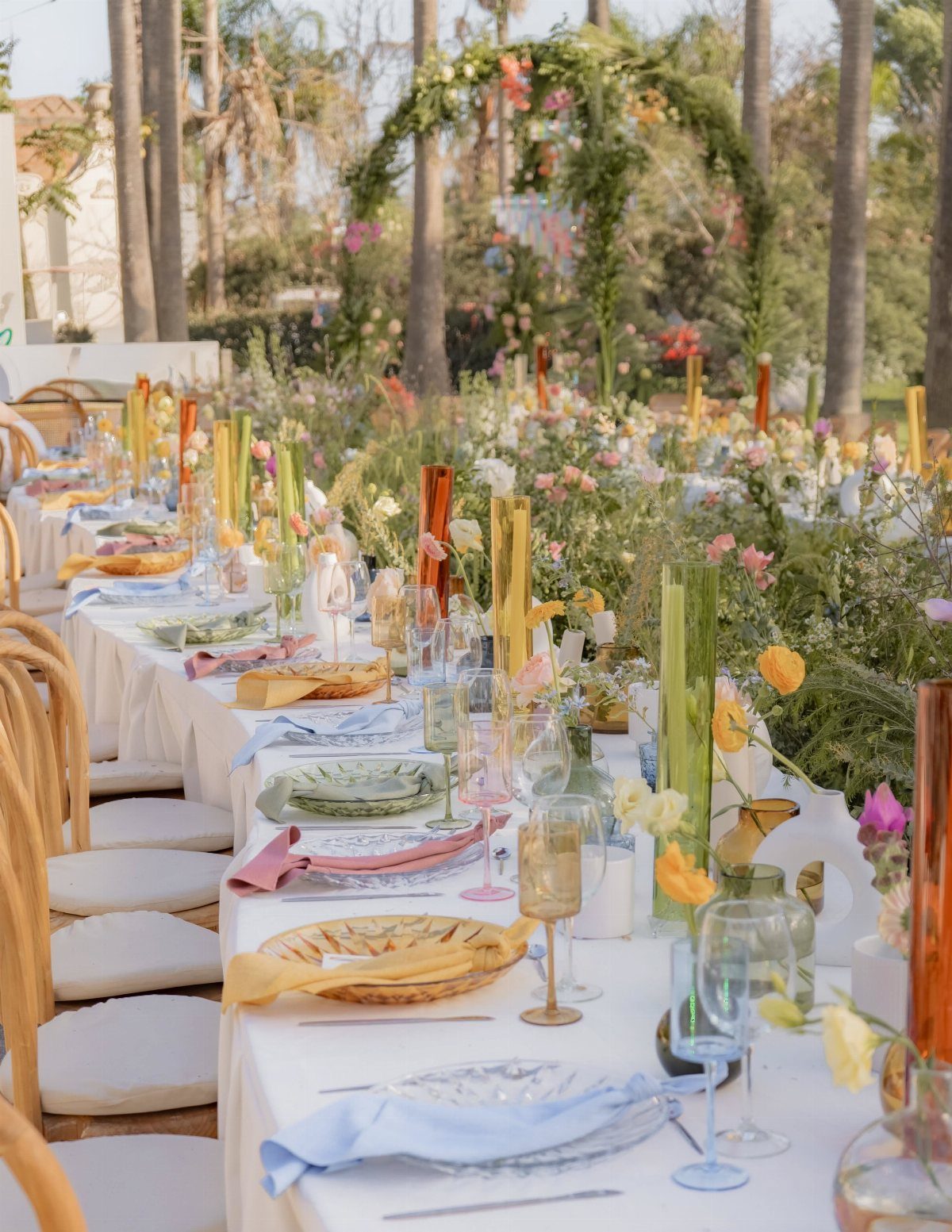 21 Colorful Wedding Table Settings for Every Style and Budget 13 A long outdoor wedding table set with colorful glassware, wildflowers, and various candles in glass holders.