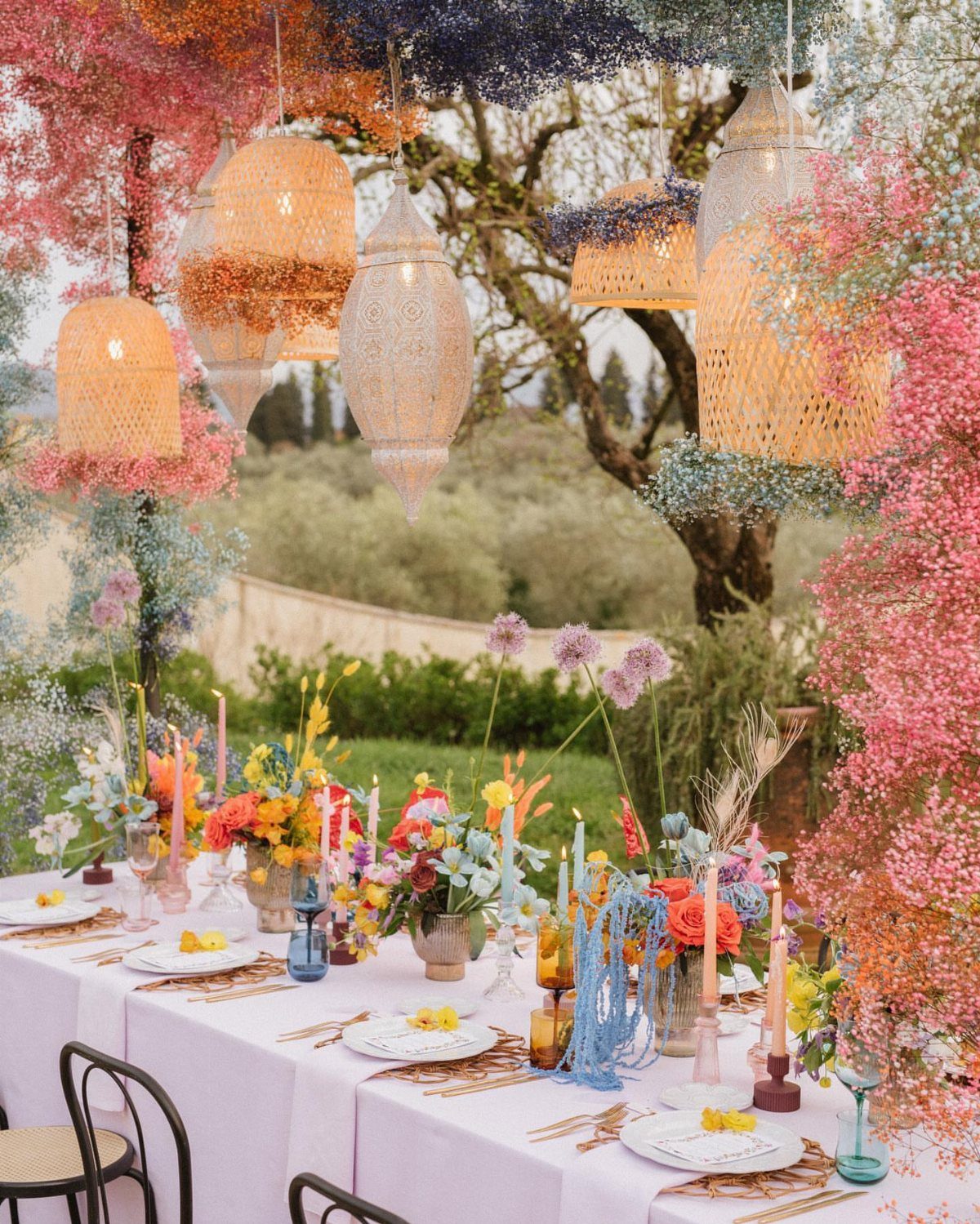 21 Colorful Wedding Table Settings for Every Style and Budget 17 A long outdoor table decorated with colorful flowers, various glassware, and hanging lanterns.