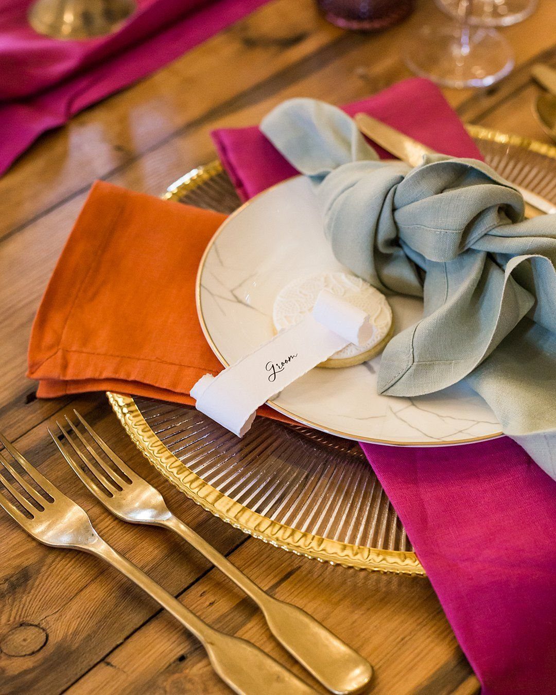 21 Colorful Wedding Table Settings for Every Style and Budget 10 Gold ribbed charger plate with white ceramic plate and colorful napkins on a wooden table.