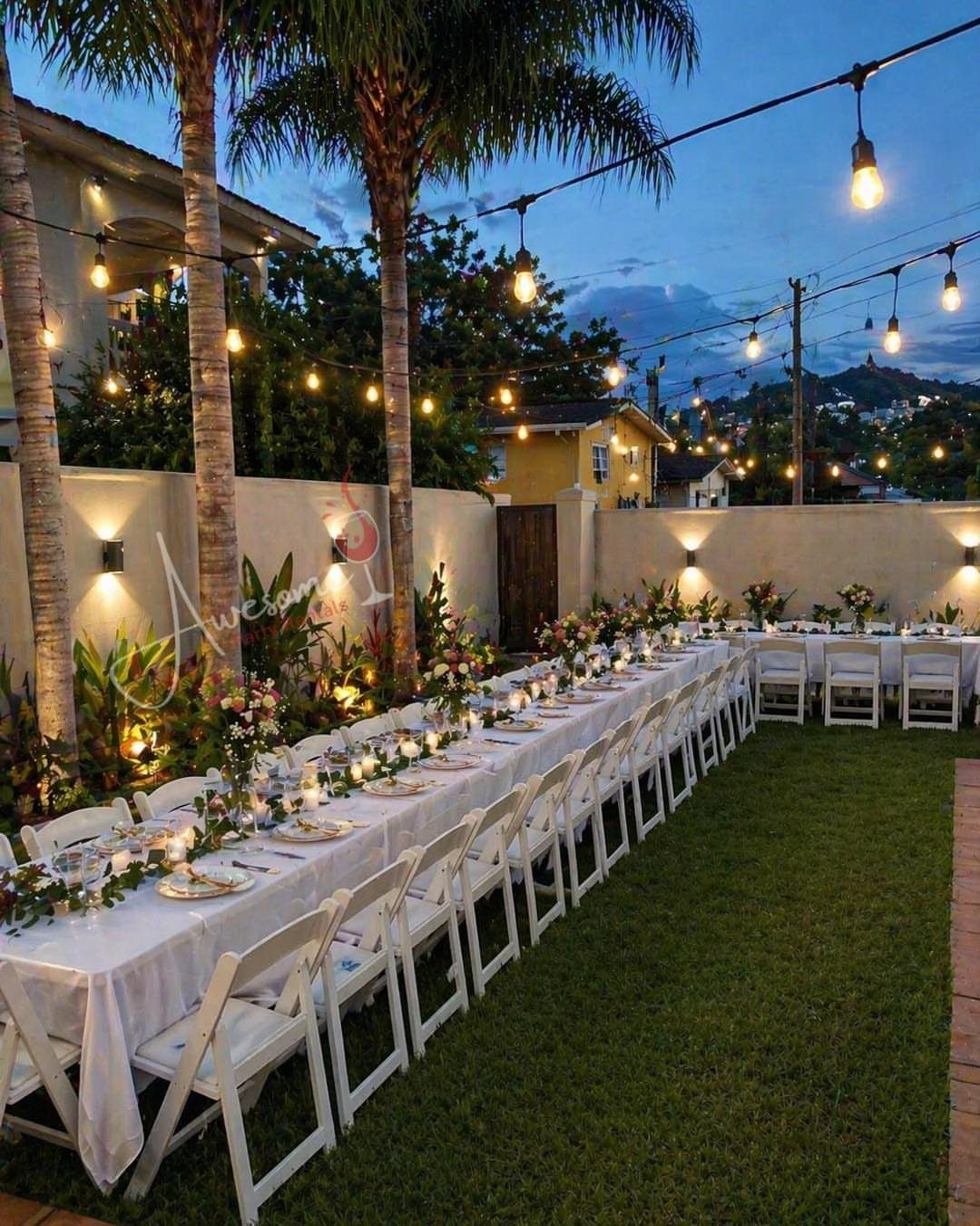 21 Colorful Wedding Table Settings for Every Style and Budget 12 Long white wedding table set with flowers and string lights on a green lawn.