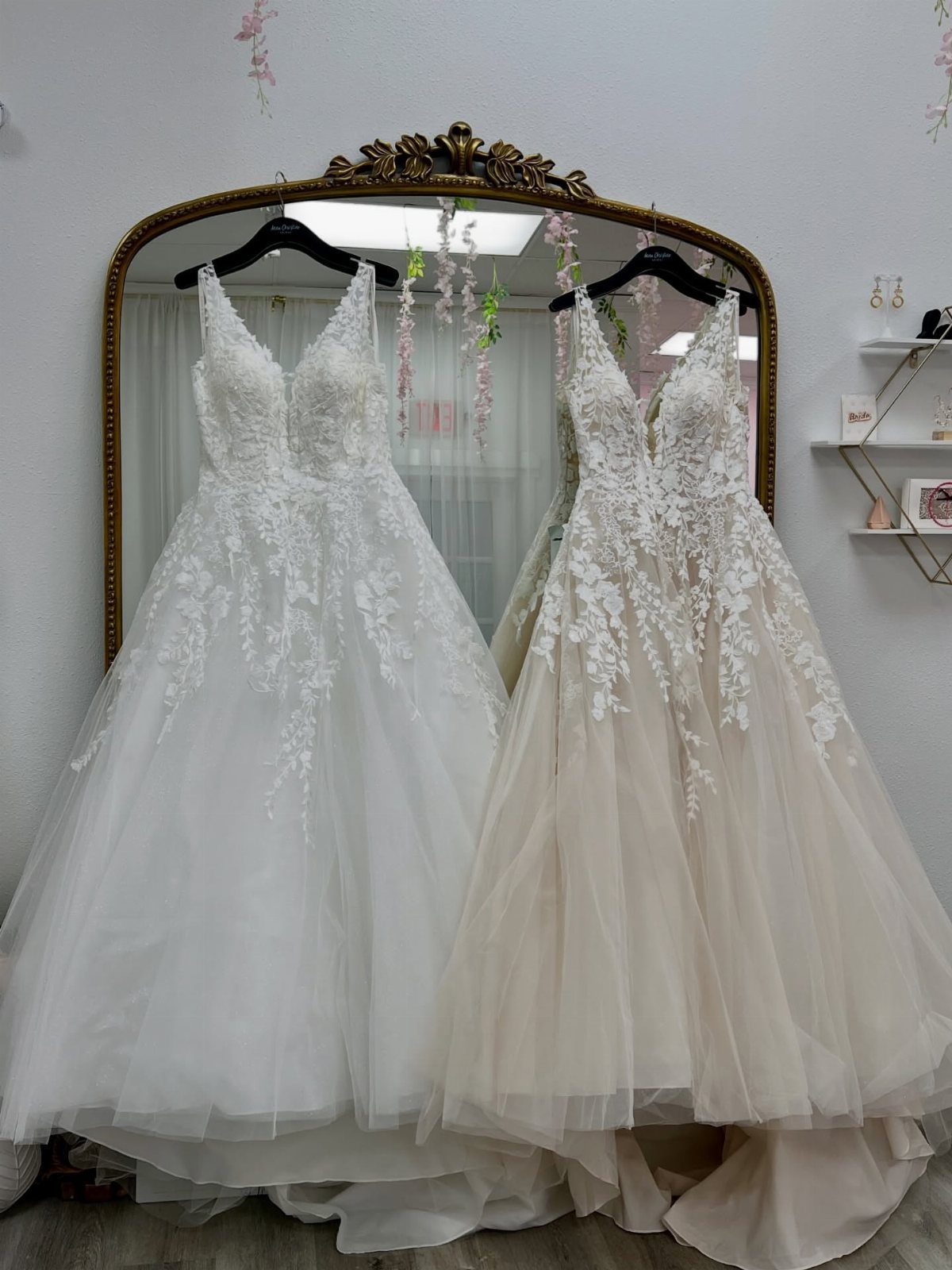 Two wedding dresses with lace bodices and tulle skirts hanging in front of a mirror.