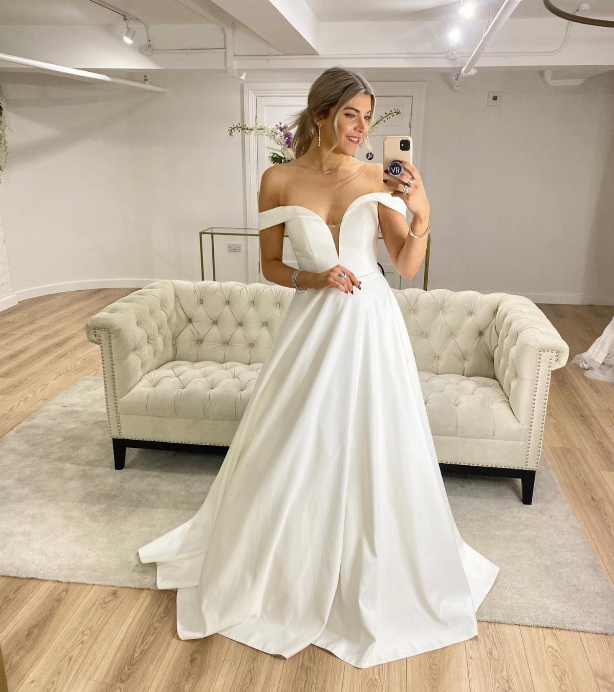 Woman in a cream white satin wedding gown posing for a mirror selfie in a bright room.