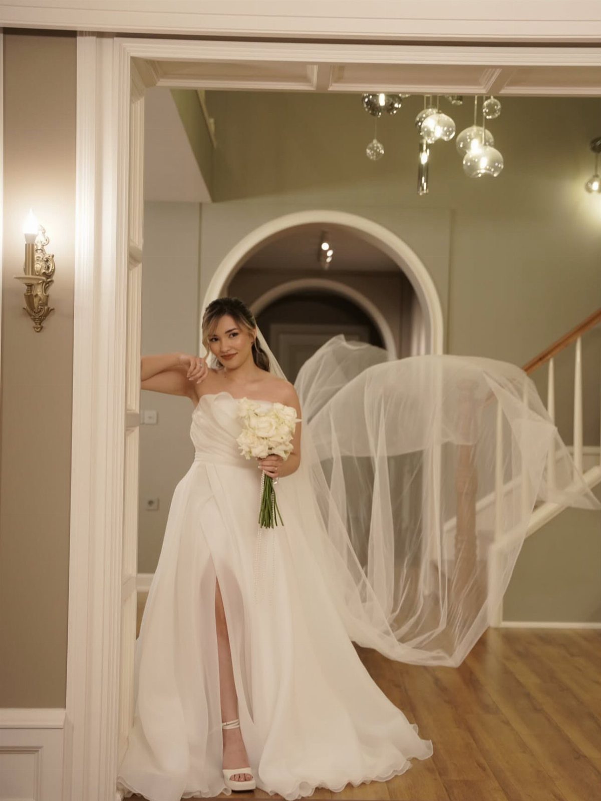 Woman in a strapless cream white wedding dress holding white roses in a doorway.