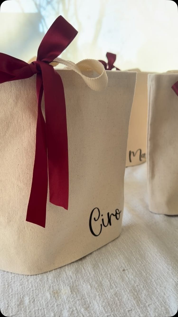 13 Cute and Fun Wedding Ideas for Guests Your Guests Will Love 3 Cream canvas tote bags with black script names and burgundy ribbon bows on a white surface.