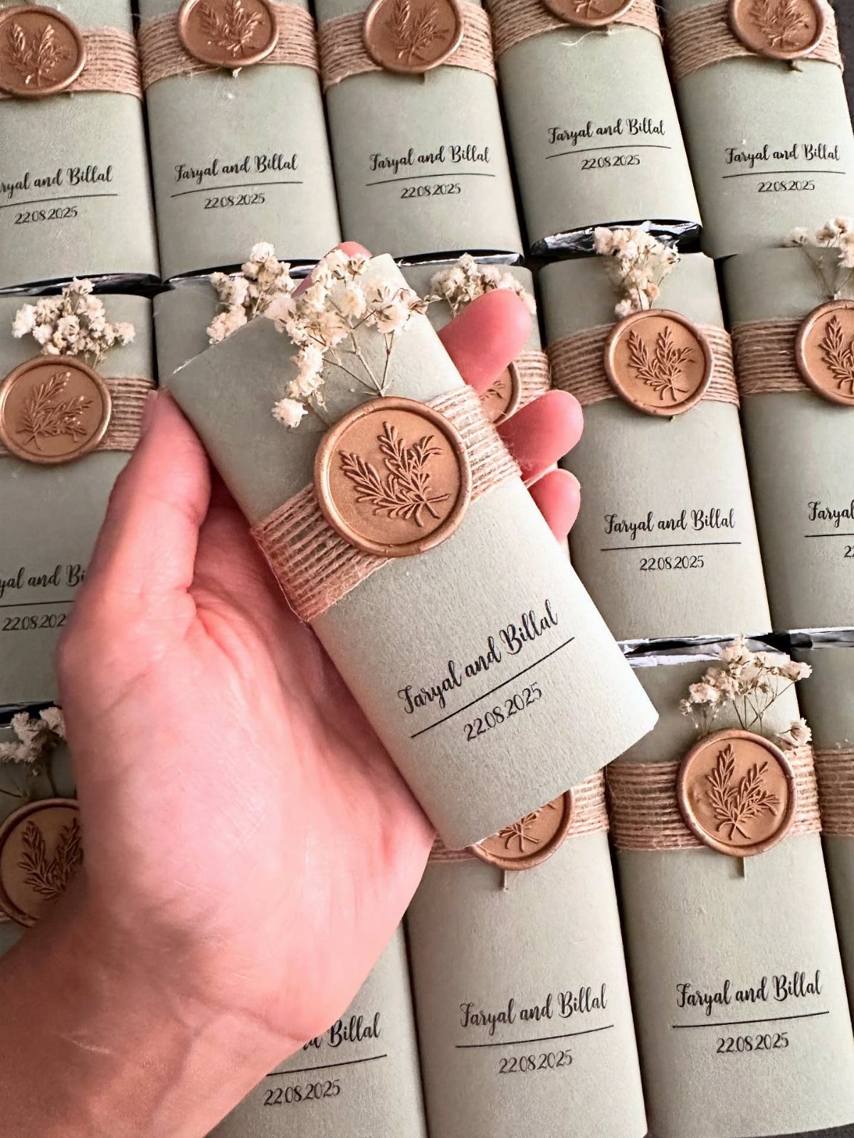 13 Cute and Fun Wedding Ideas for Guests Your Guests Will Love 9 A hand holding a grey cylindrical wedding favor wrapped in twine with a wax seal and dried flowers.