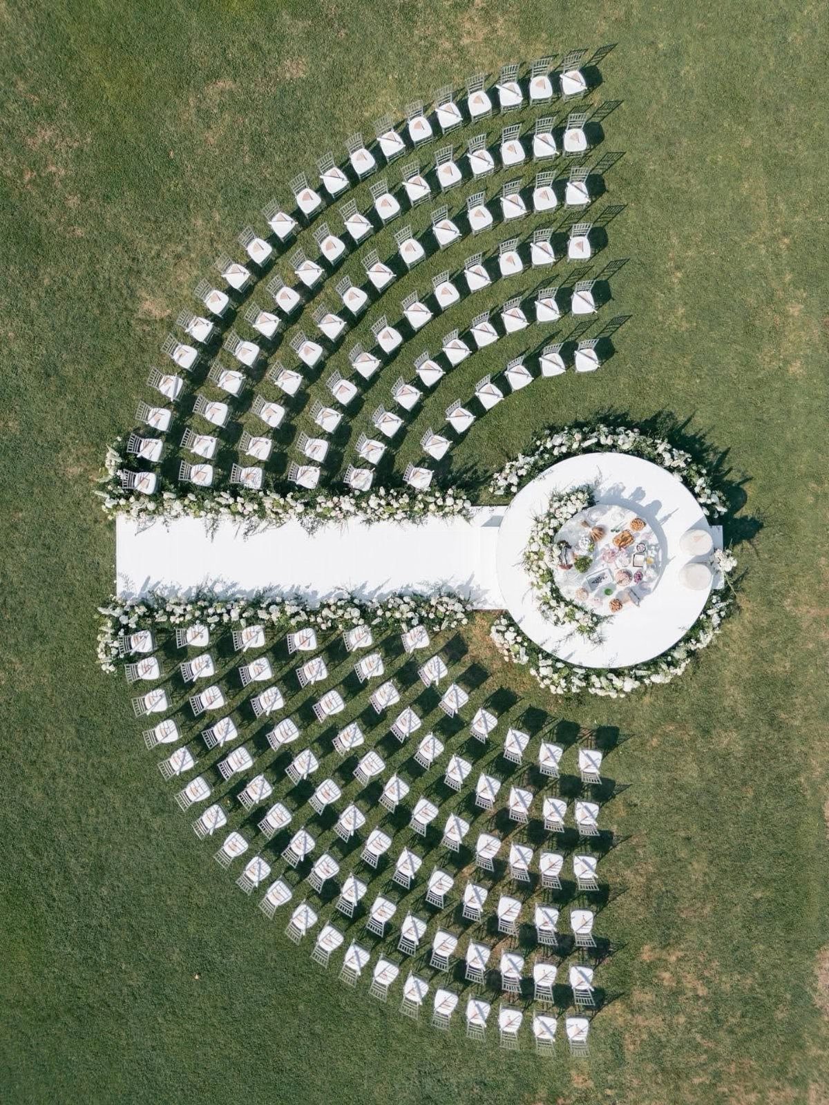 Aerial view of white wedding chairs arranged in semi-circles on a green lawn around a floral altar.