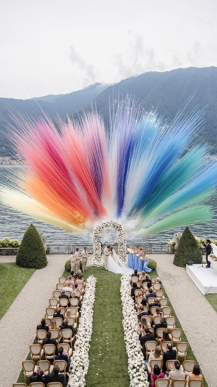 A wedding ceremony set outdoors by a lake with a large rainbow-colored burst behind the arch.
