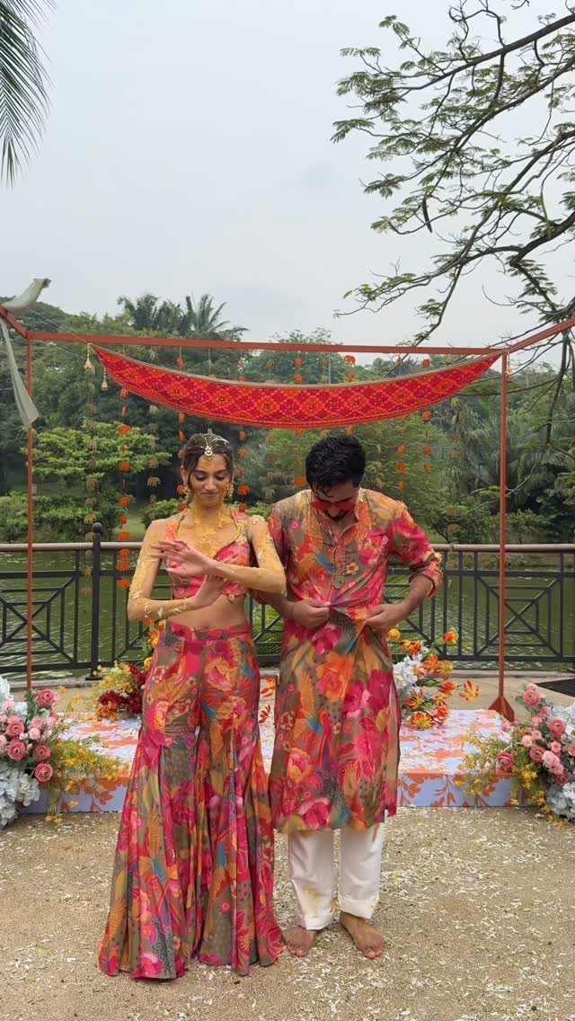 A couple wearing matching vibrant floral patterned outfits standing under a red canopy outdoors.