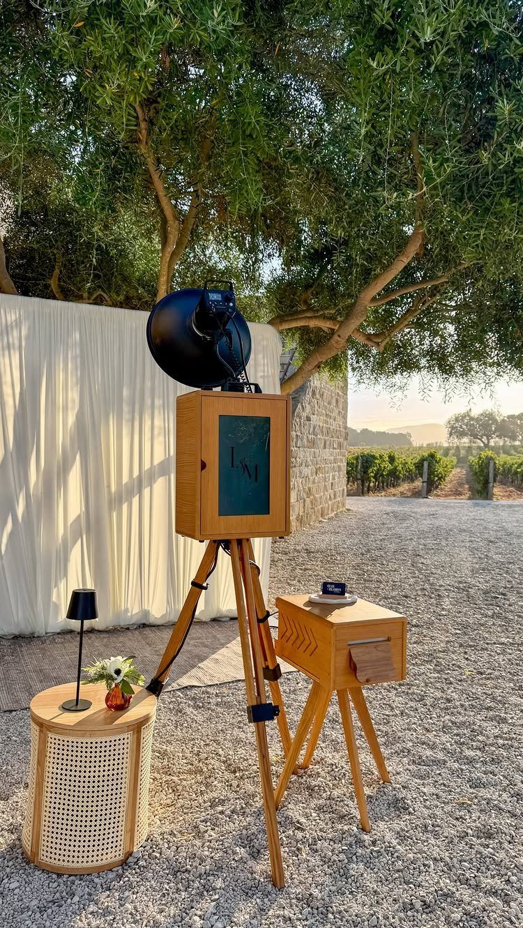 A vintage wooden photo booth on a tripod stands on gravel near trees.