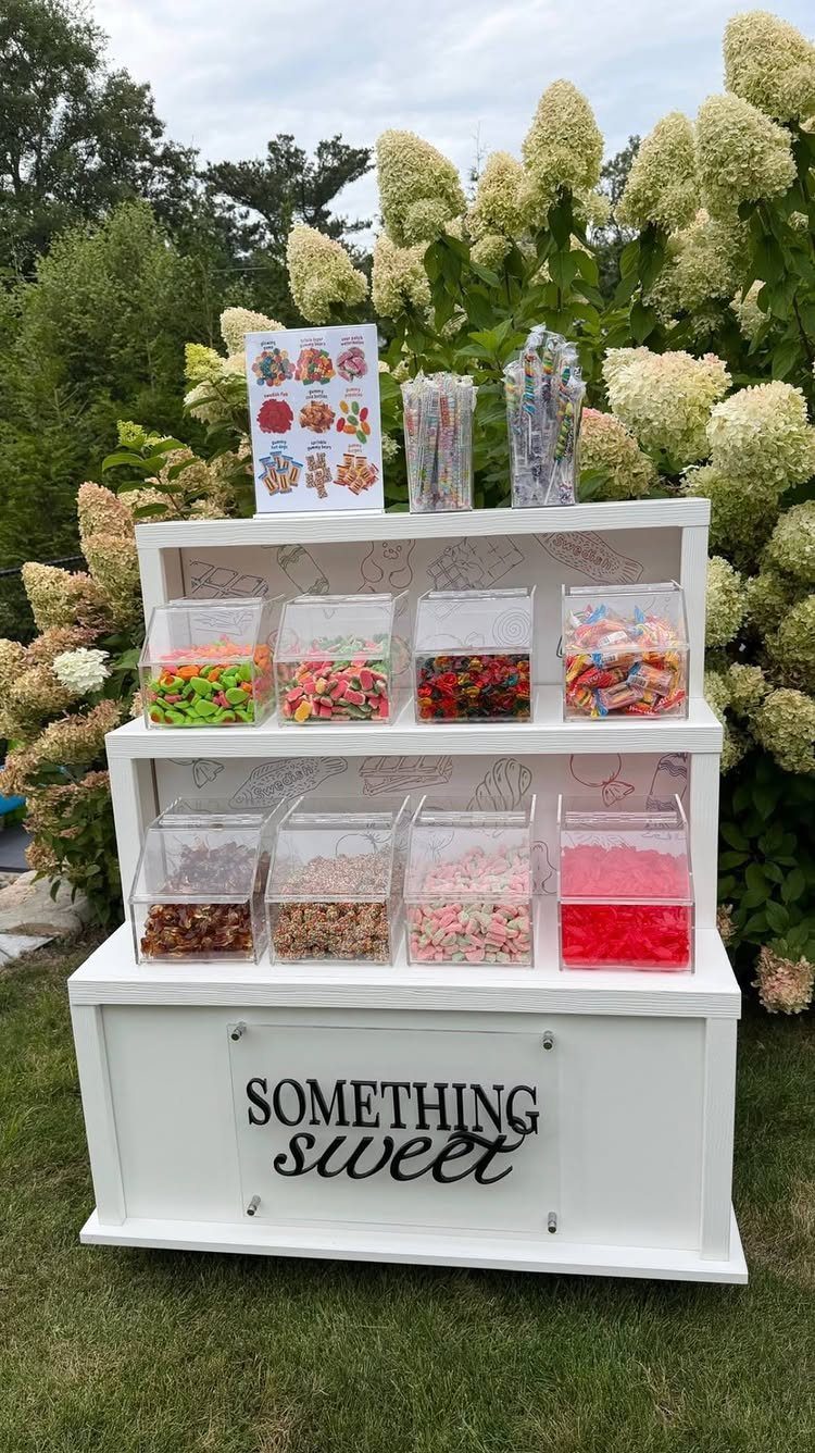 A white tiered wooden stand filled with various colorful candies in clear containers outdoors.