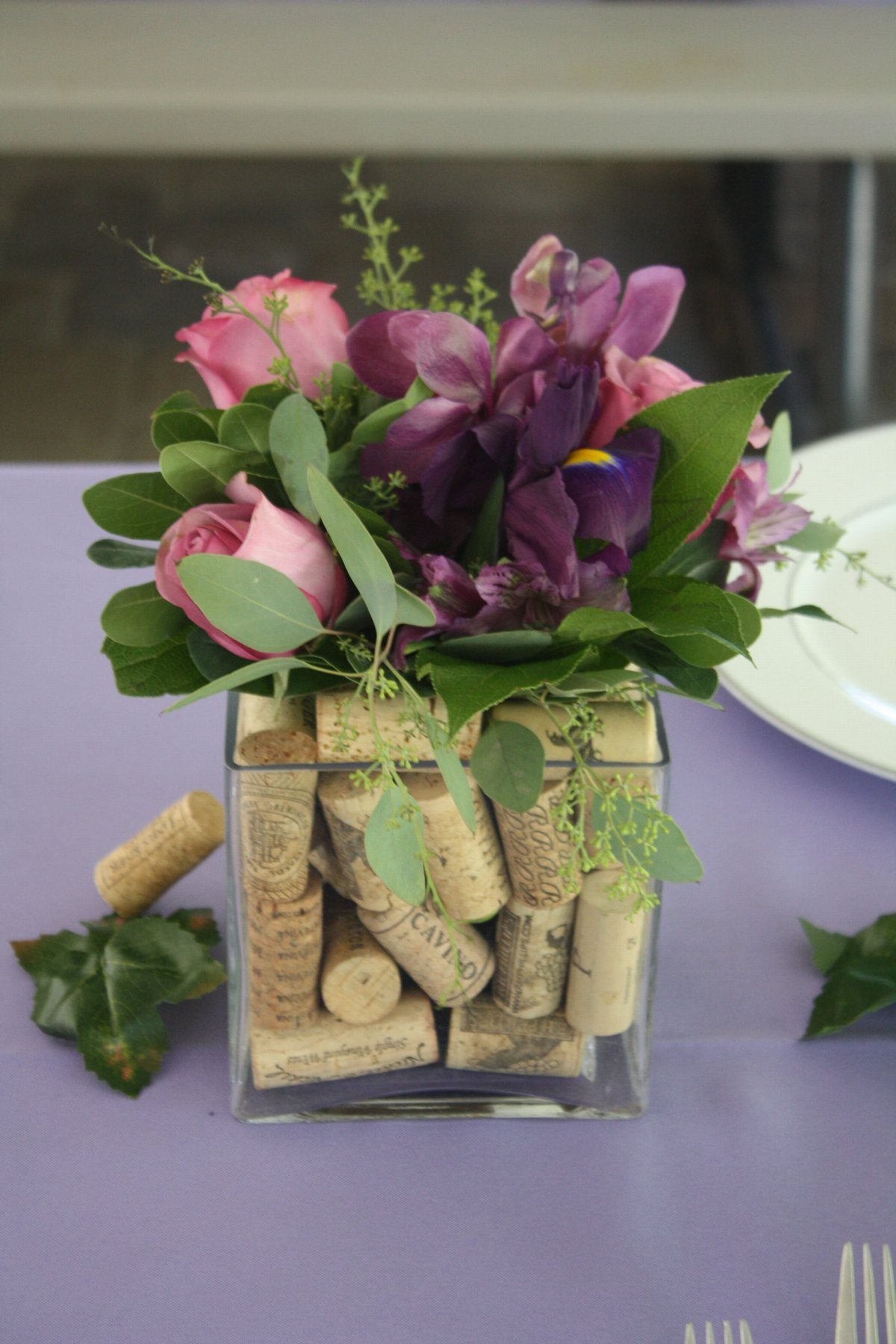 8 Dollar Tree Wedding Decor 3 Pink roses and purple irises arranged in a glass vase filled with wine corks on a lavender table.