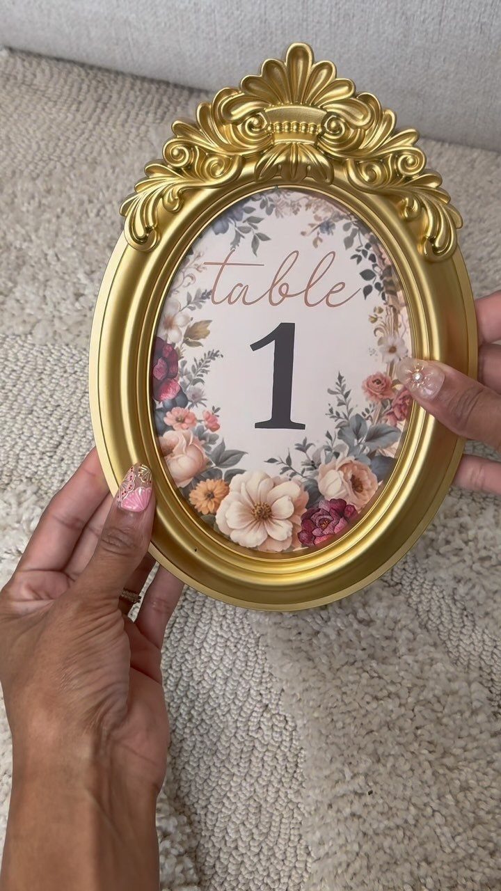 8 Dollar Tree Wedding Decor 5 Hands holding an ornate gold oval frame with a floral table number one print.