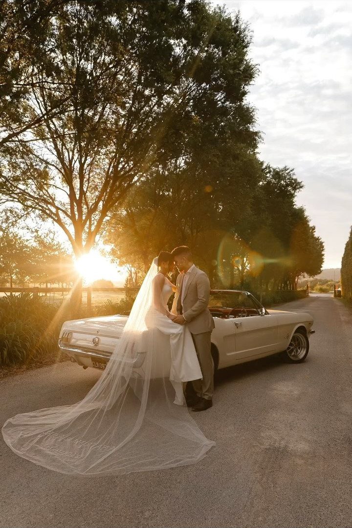 10 Ethereal and Romantic Wedding Ideas That Feel Like a Dream 6 A bride and groom pose by a vintage car during a golden hour sunset.
