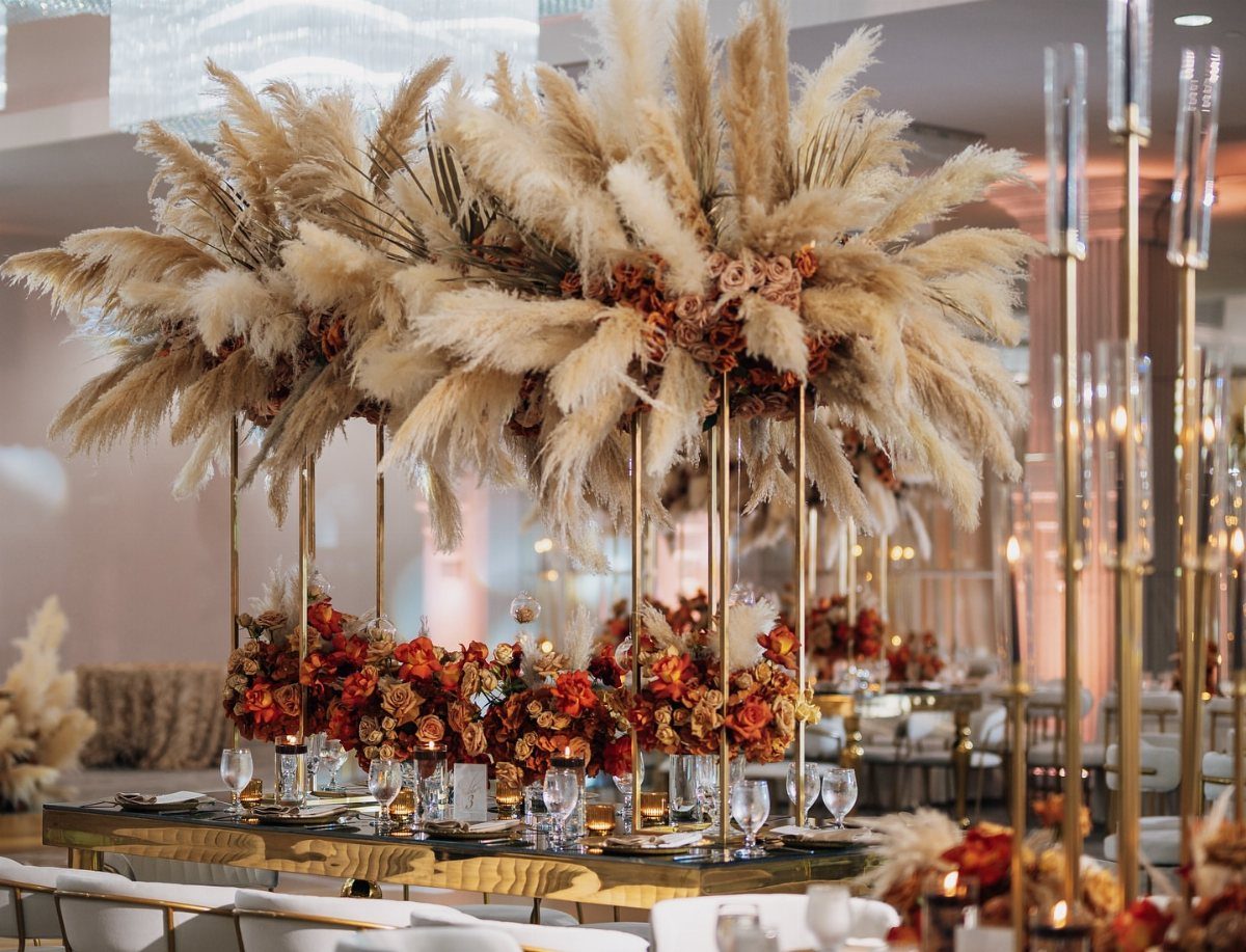 Tall gold metal stands holding large arrangements of pampas grass and autumn flowers on a table.