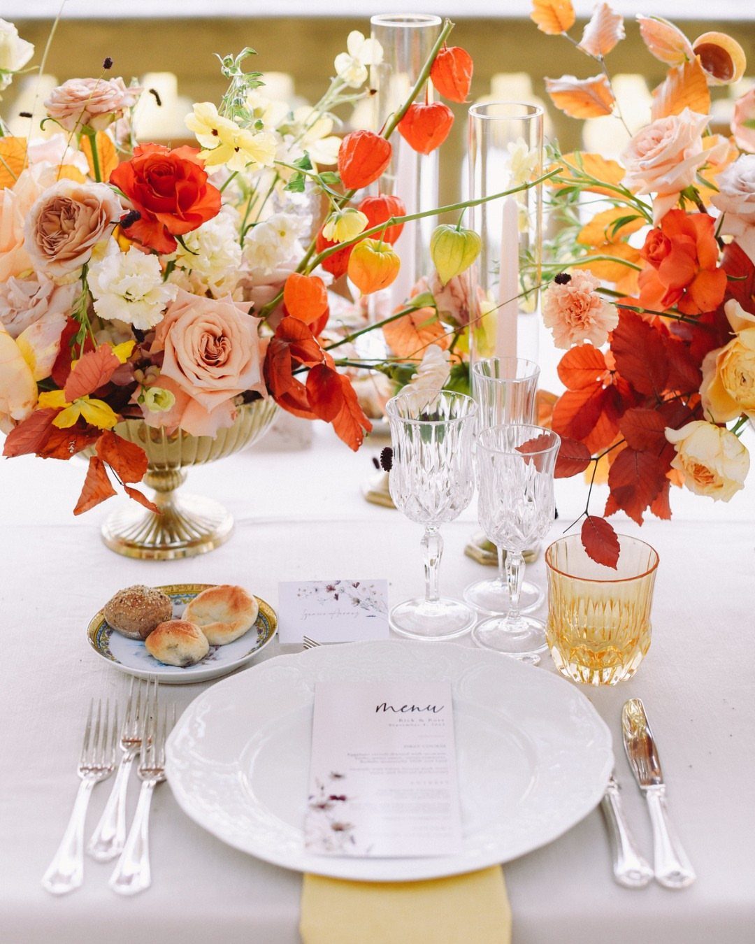 Floral centerpieces with orange and peach roses arranged in gold bowls on a formal dining table.