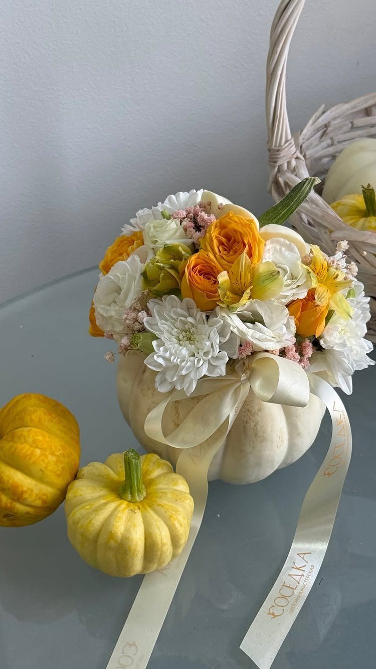 White pumpkin filled with yellow and white autumn flowers sitting next to small yellow gourds.