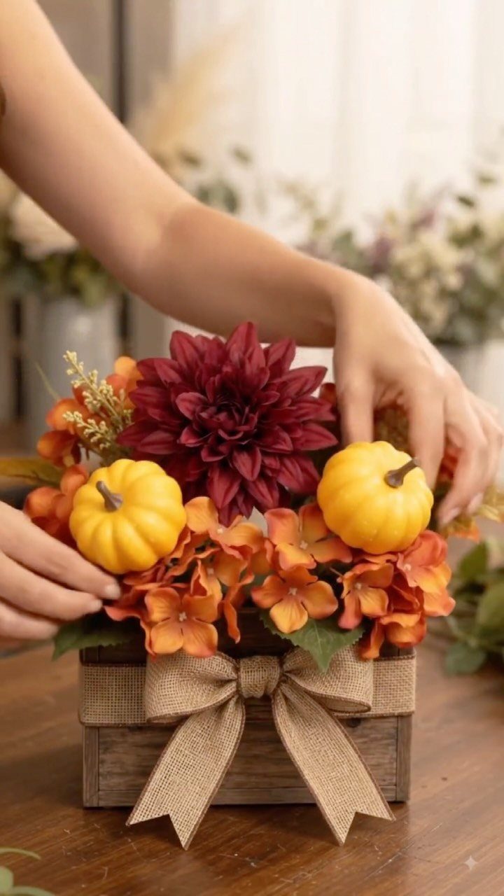 Hands arranging a fall floral centerpiece with burgundy dahlias, orange hydrangeas, and small pumpkins in a wooden box.