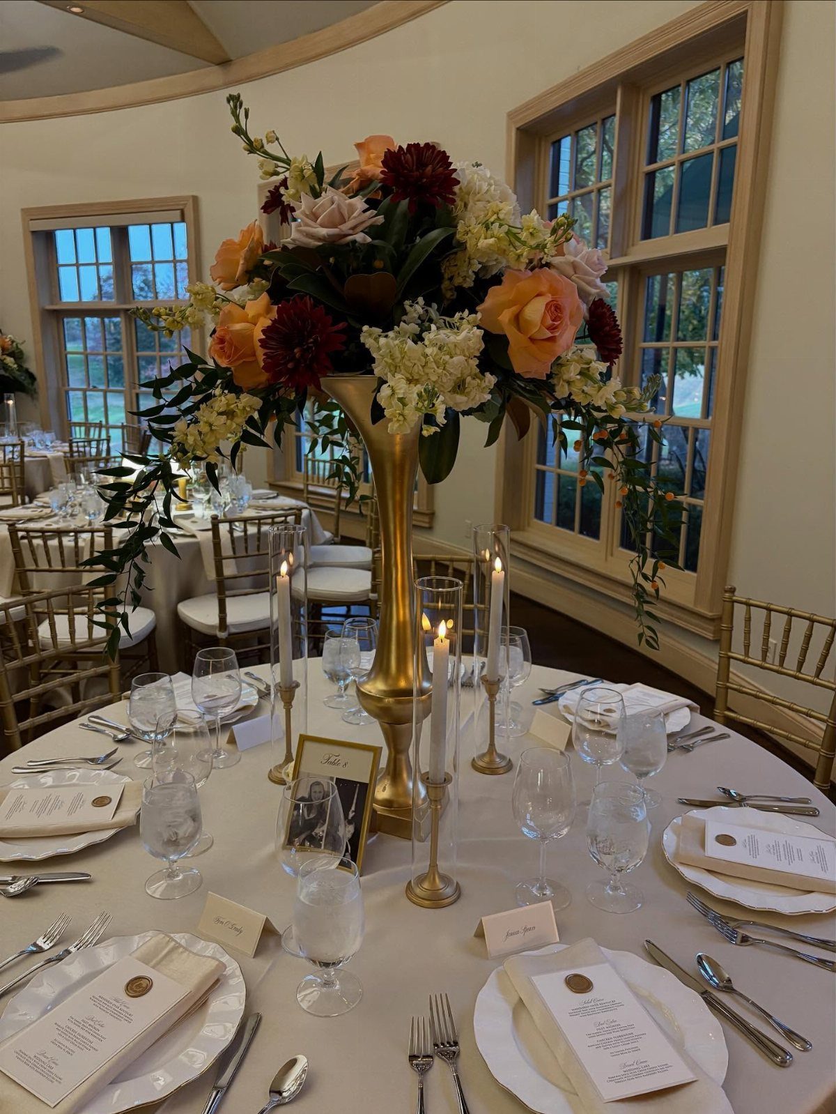A tall gold vase filled with autumn-colored flowers serves as a wedding centerpiece on a set table.