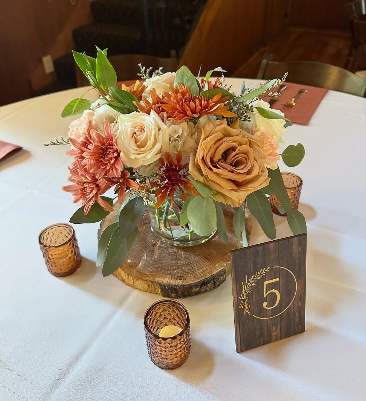 Floral centerpiece with orange dahlias and cream roses on a wooden slice with amber votive candles.