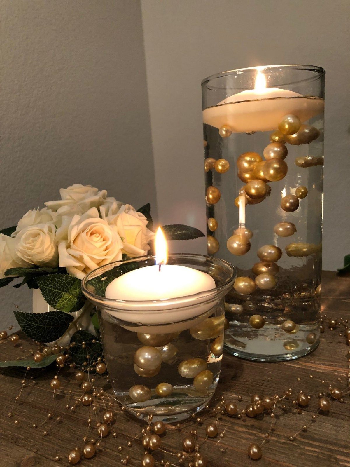 Gold pearls floating in water inside glass vases with white floating candles and roses.