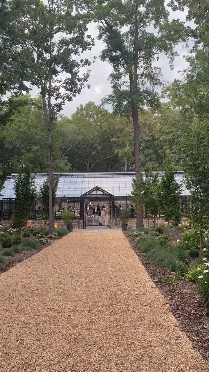 Garden Estate Wedding: 12 Ideas for Ceremony and Reception 8 A gravel walkway leading toward a large glass greenhouse surrounded by tall green trees and garden plants.