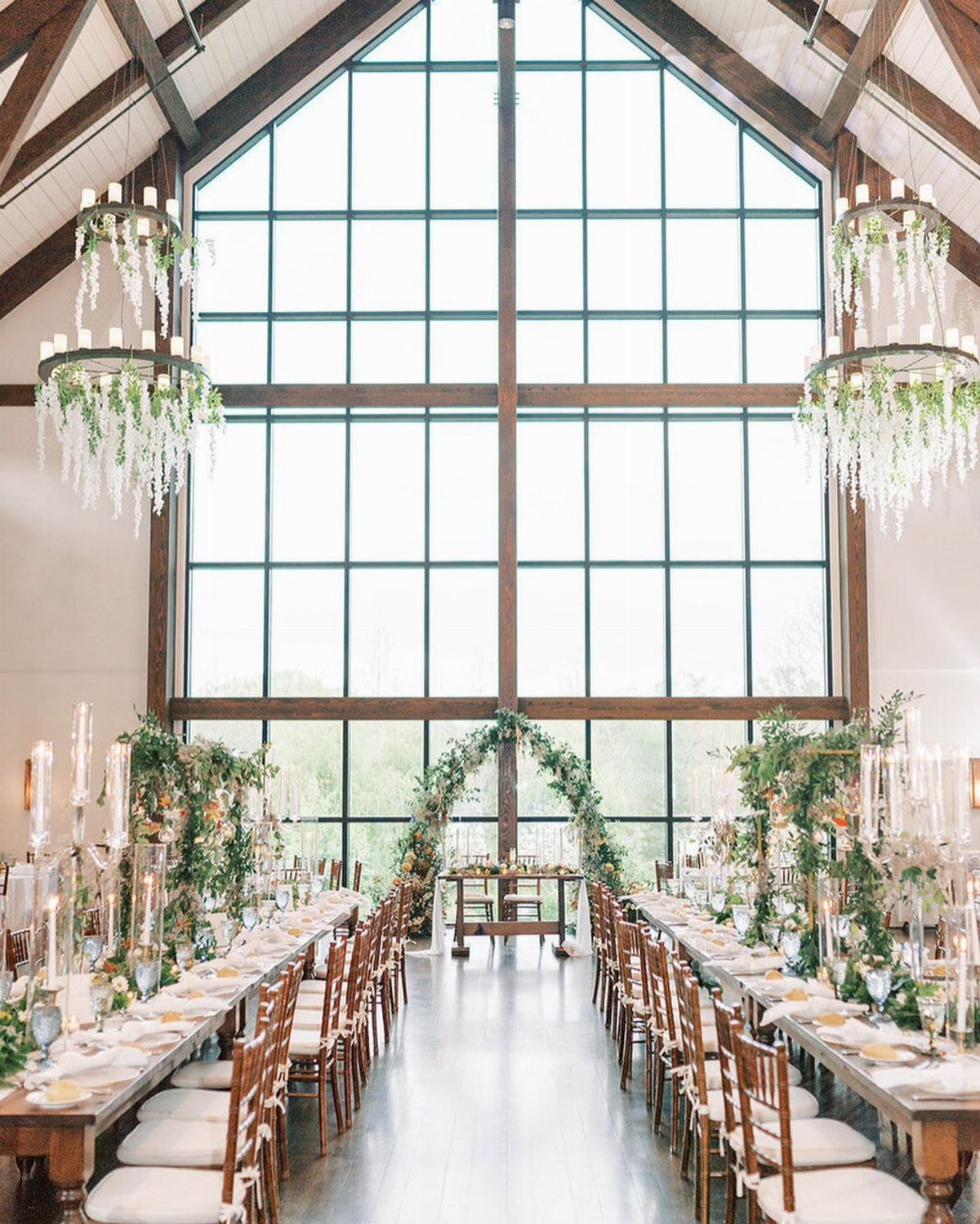 Garden Estate Wedding: 12 Ideas for Ceremony and Reception 9 Long wooden wedding banquet tables decorated with greenery and candles inside a bright, high-ceilinged estate venue.