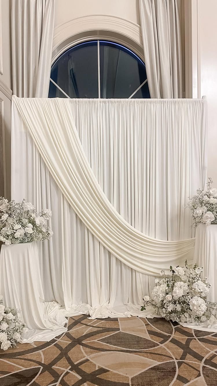 23 Indoor Wedding Decorations for Ceremony and Reception 8 White fabric backdrop with draped cream fabric and white floral arrangements on pedestals.