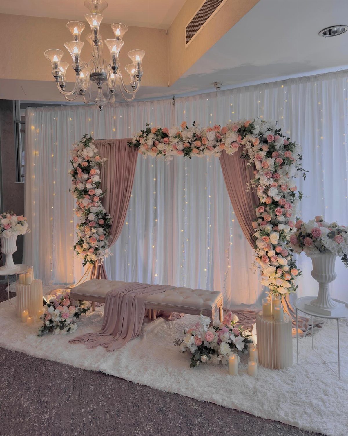 23 Indoor Wedding Decorations for Ceremony and Reception 9 Floral arrangements decorating a pink fabric wedding arch against white fairy light curtains.