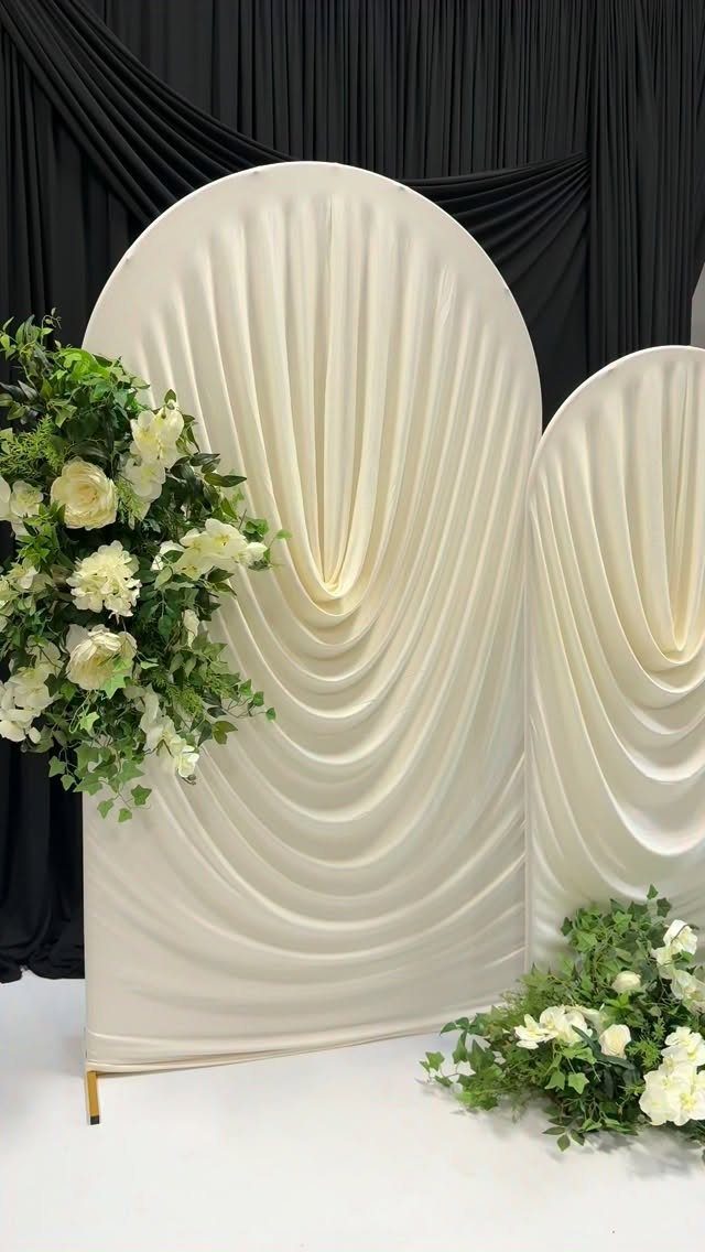 23 Indoor Wedding Decorations for Ceremony and Reception 22 Cream draped fabric arches decorated with white flowers against a black curtain backdrop.