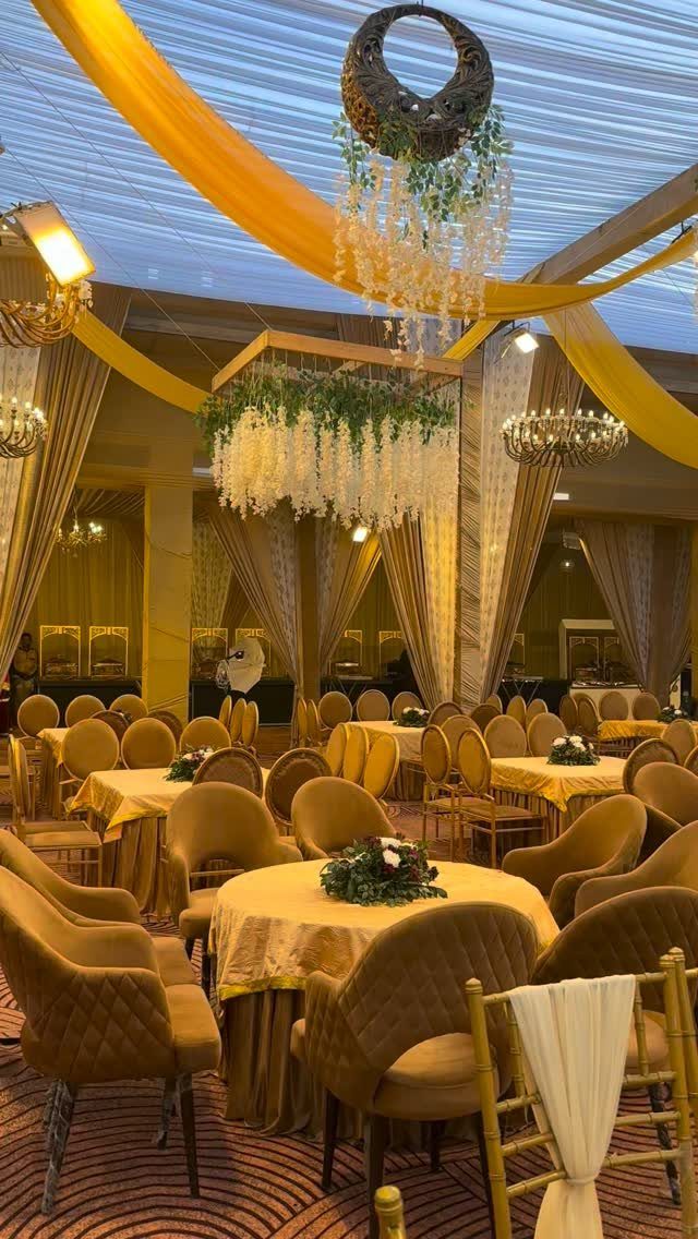 23 Indoor Wedding Decorations for Ceremony and Reception 12 Round banquet tables with gold linens and tan velvet chairs under yellow ceiling draping.