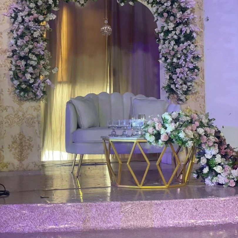 23 Indoor Wedding Decorations for Ceremony and Reception 6 A light blue scalloped loveseat and gold geometric table set against gold curtains and floral arches.