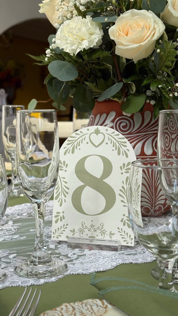 10 Lace and Greenery Wedding Table Ideas to Try 8 A wedding table setting with white roses, greenery, glassware, and a table number eight.