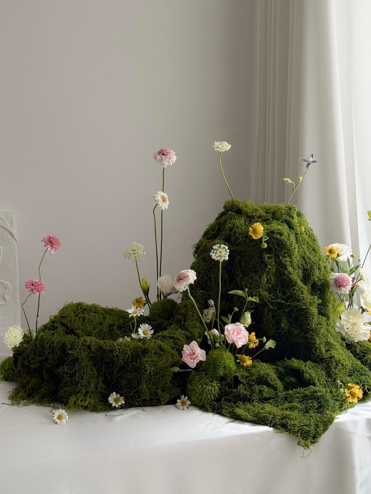 10 Lace and Greenery Wedding Table Ideas to Try 3 Small pink and white wildflowers growing out of a mound of green moss on a white surface.