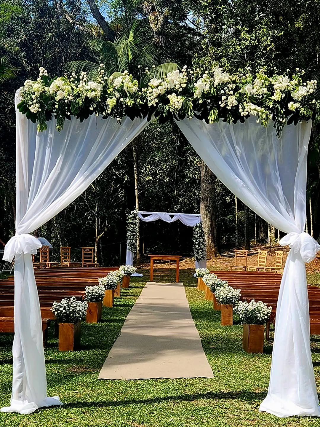 White floral wedding arch and aisle runner lined with flower boxes in a forest setting.
