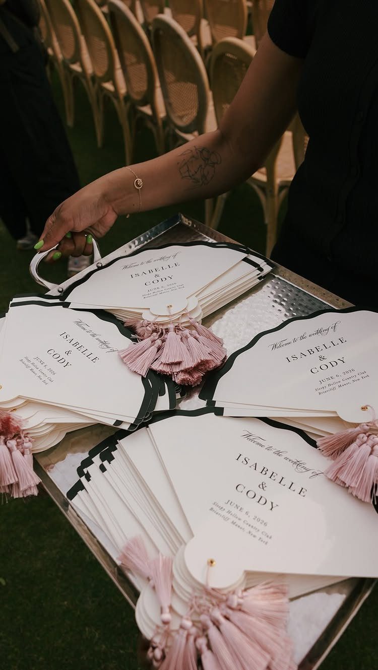 A silver tray holding white paper wedding ceremony program fans with pink tassels.