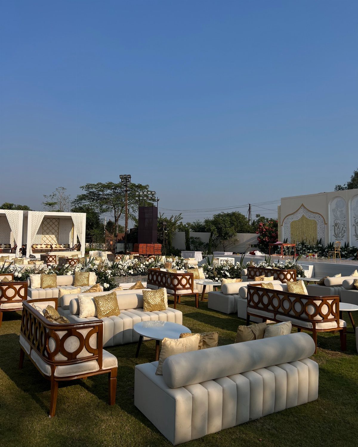 White sofas and wooden armchairs with gold cushions arranged on a green lawn outdoors.
