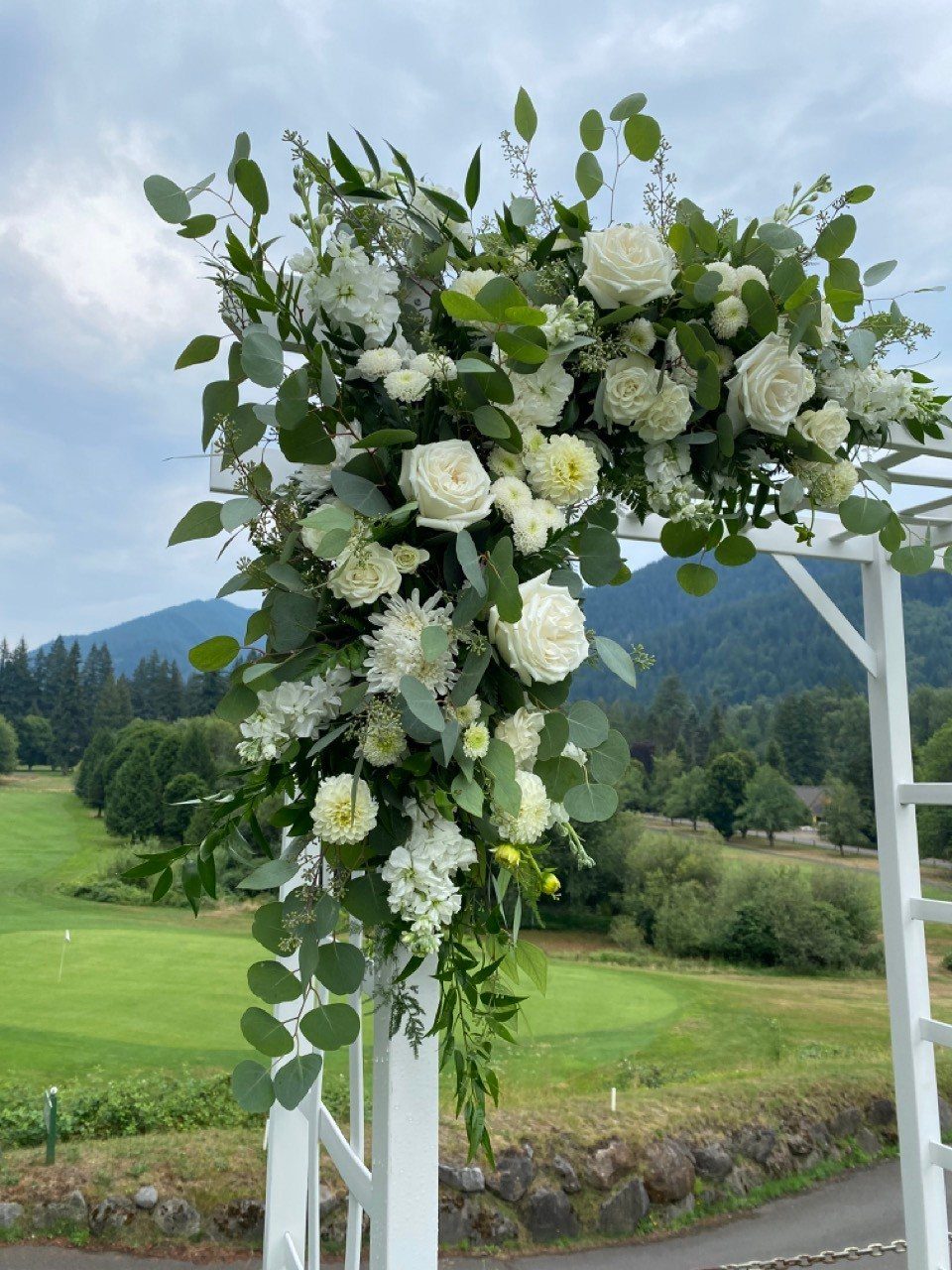 White roses and green eucalyptus leaves decorating the corner of a white wooden wedding arch outdoors.