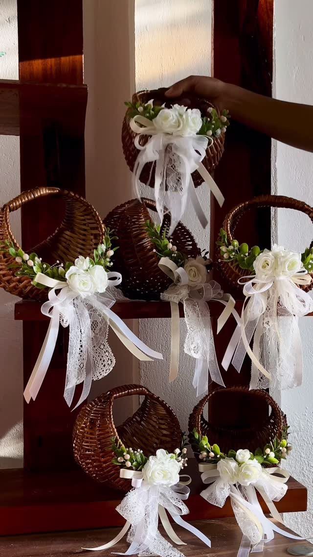 Small wicker baskets decorated with white roses and lace ribbons sitting on a wooden shelf.