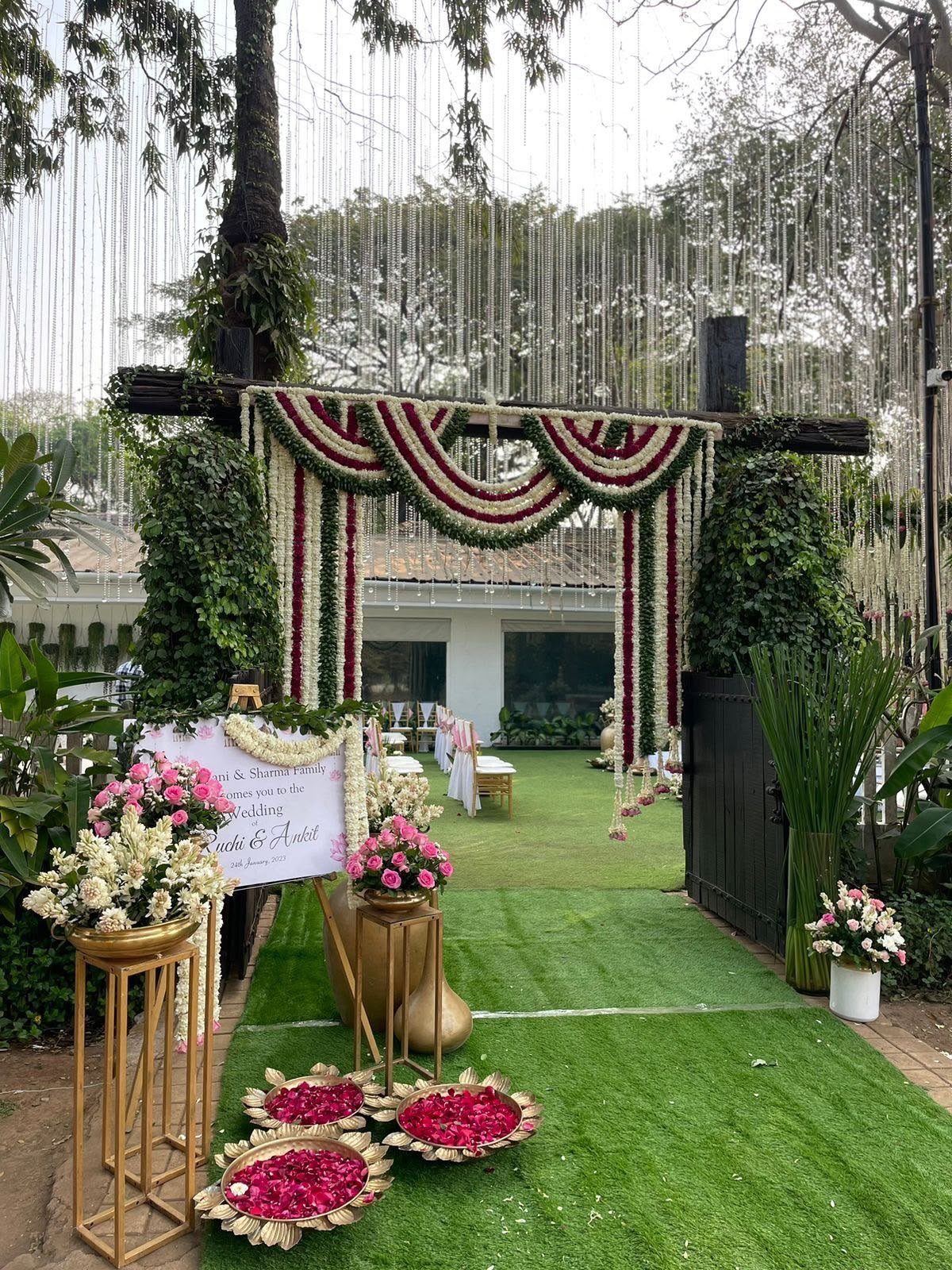 A wooden wedding arch decorated with thick red, white, and green flower garlands over a green lawn.