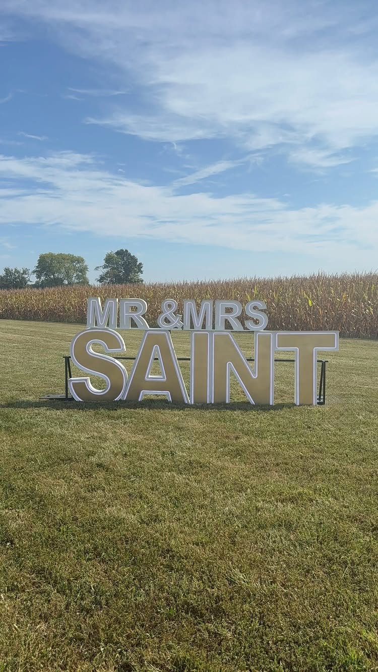 Large illuminated marquee letters spelling MR & MRS SAINT on a green grass field.